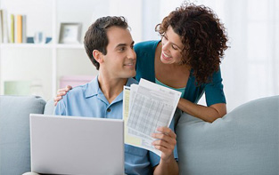 Young woman leaning over her husband while he reviews budget documents and uses the online Debt Management tool.